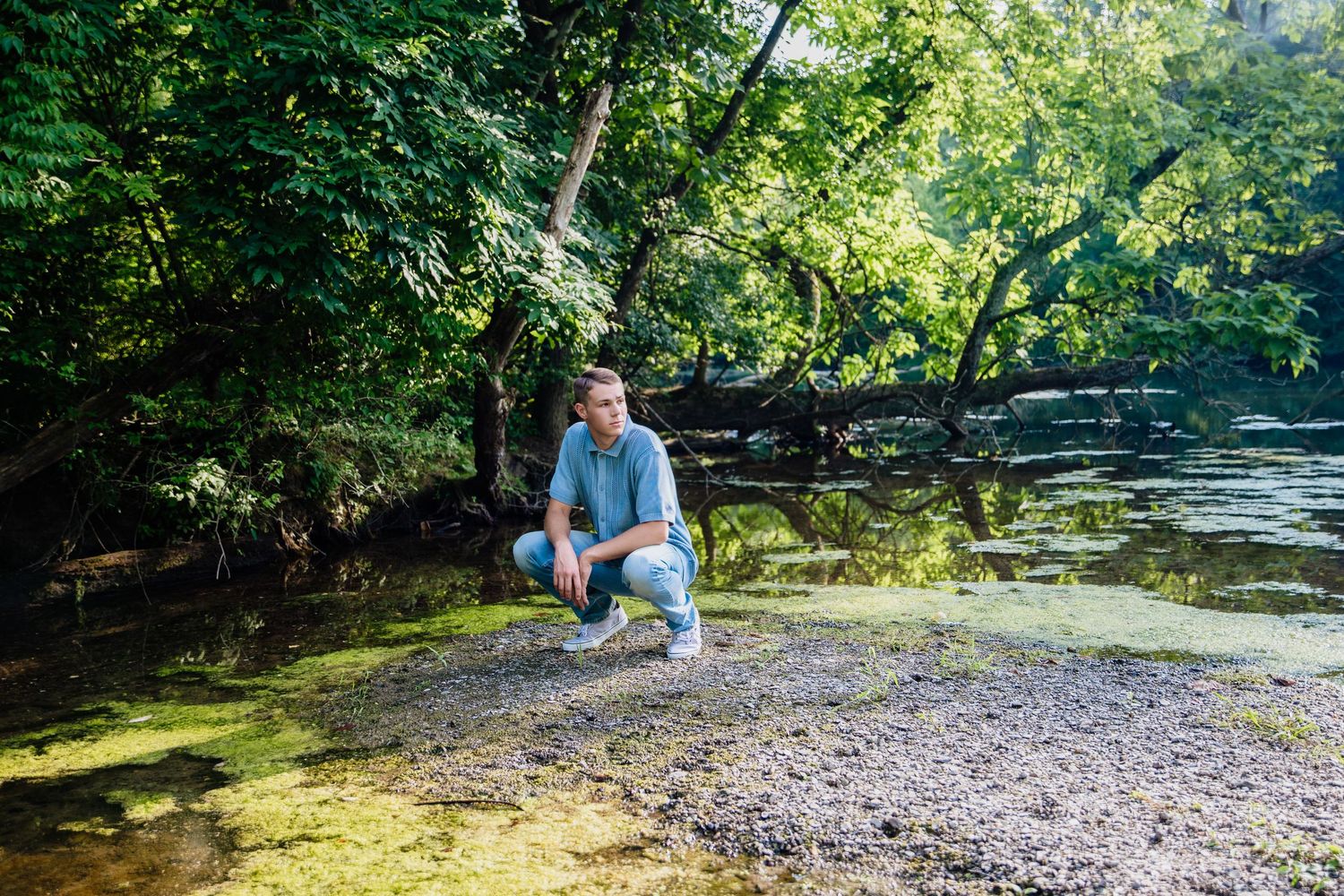 Young man crouching by a serene river bank surrounded by lush green trees on a sunny day.