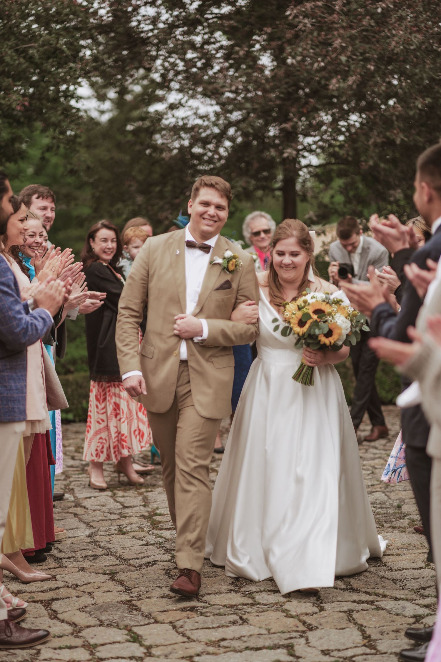 Smiling bride and groom walk arm in arm down a cobblestone path as wedding guests applaud, the bride holding a sunflower bouquet.