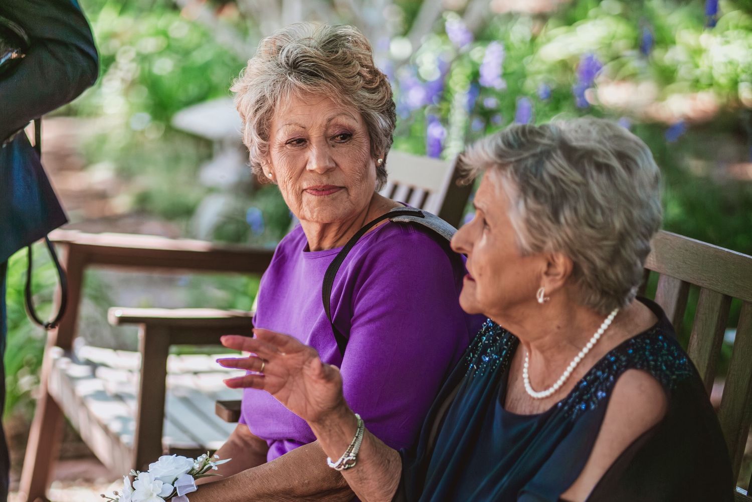 Two older women sit side by side on a wooden garden bench, talking and gesturing while surrounded by lush greenery and purple flowers.