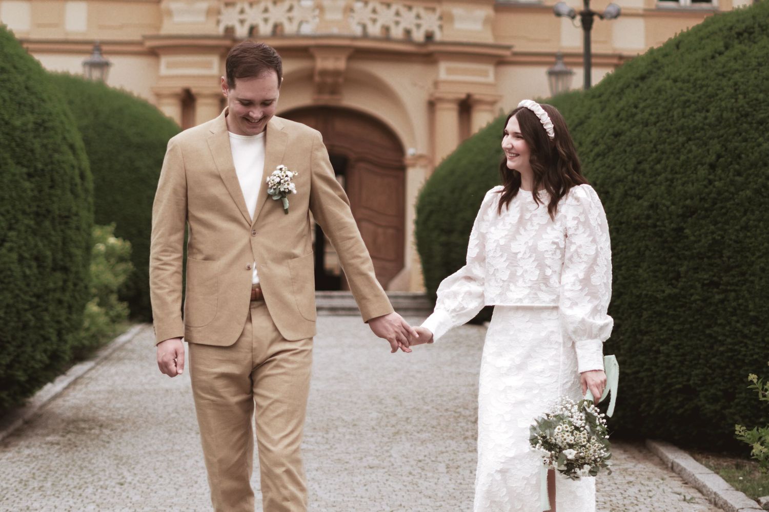 Smiling bride and groom in a tan suit hold hands while walking down a gravel path near hedges and a historic building.