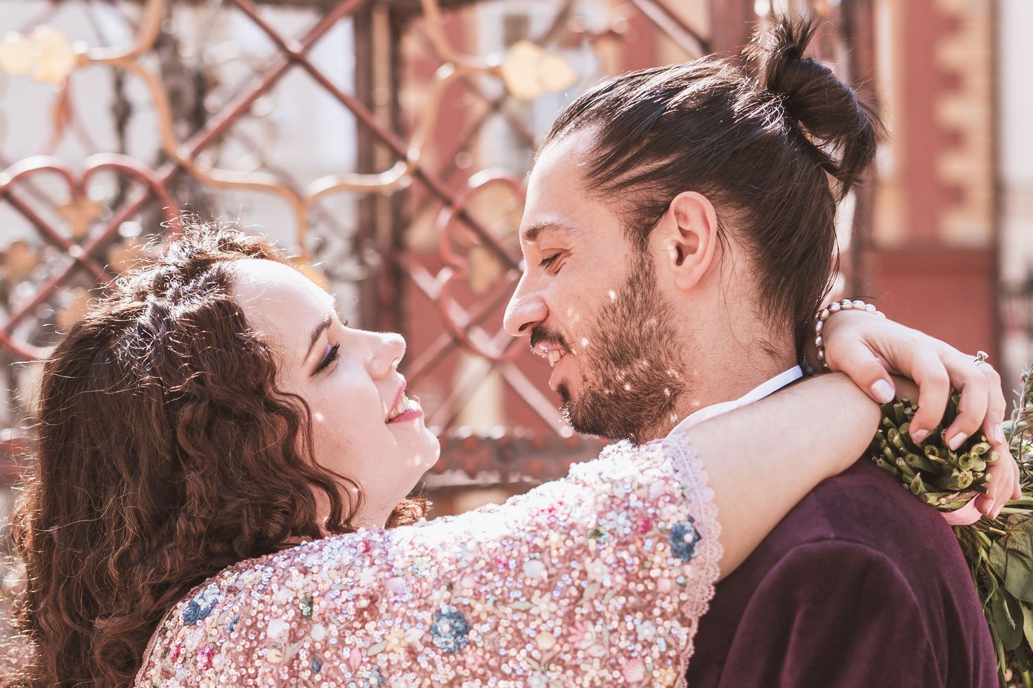 A smiling woman with curly brown hair in a sequined floral top embraces a bearded man with a bun in front of an ornate metal gate.