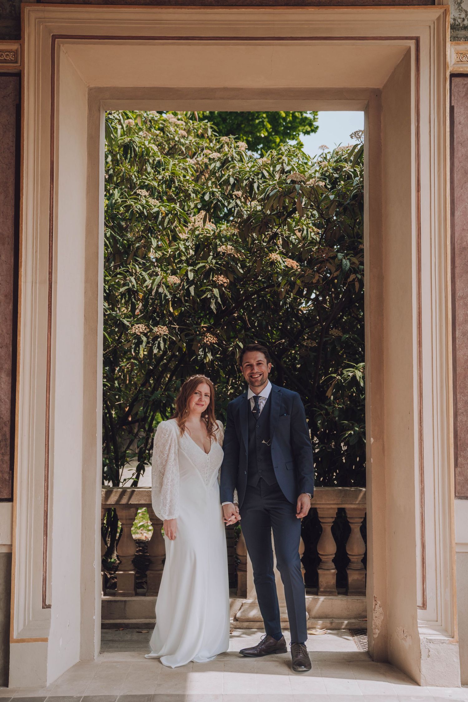 Bride in a white long-sleeve gown and groom in a navy suit hold hands in a stone archway framed by green trees.