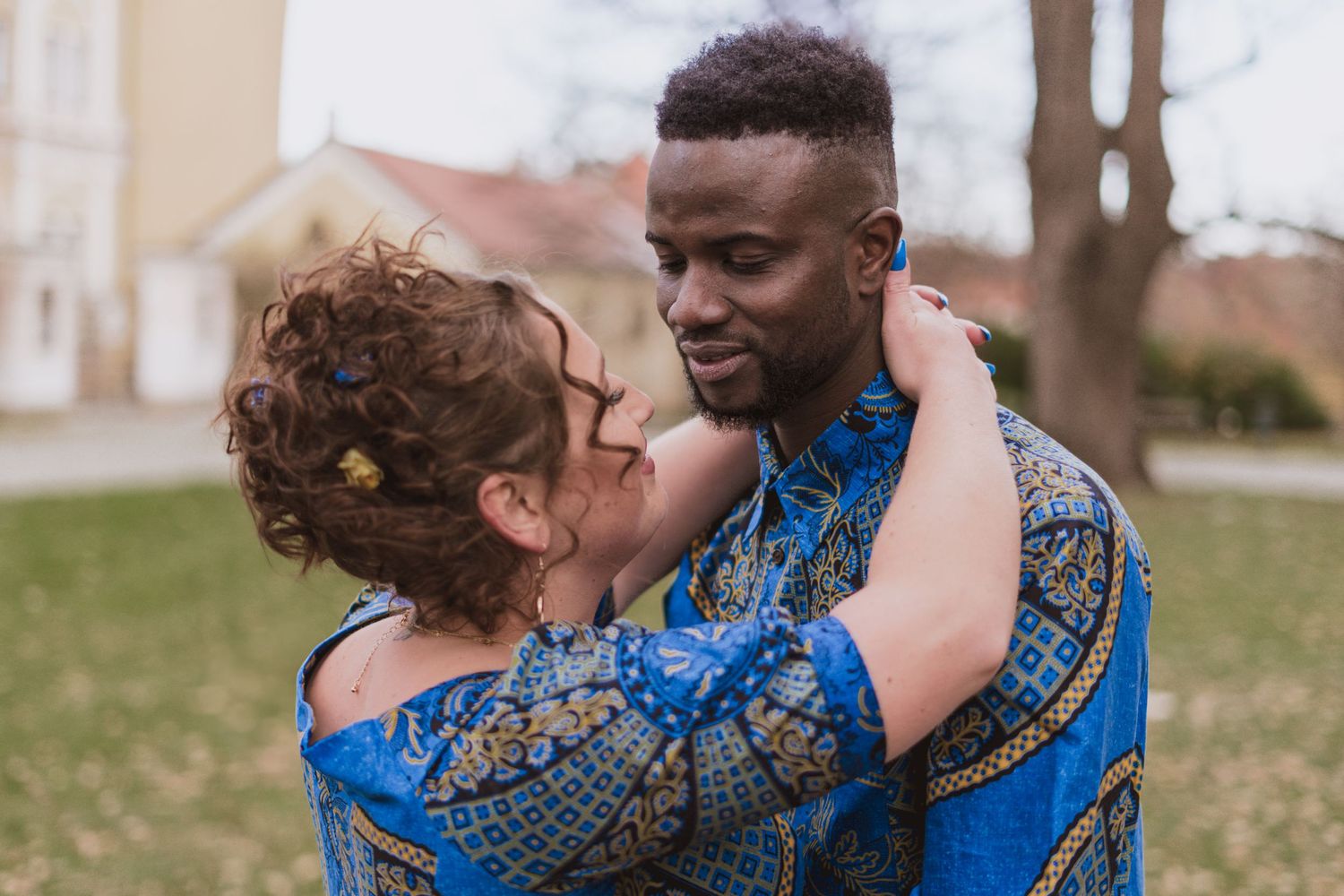 Smiling couple in matching blue African print shirts embrace outdoors in a park with bare trees and a house in the background.