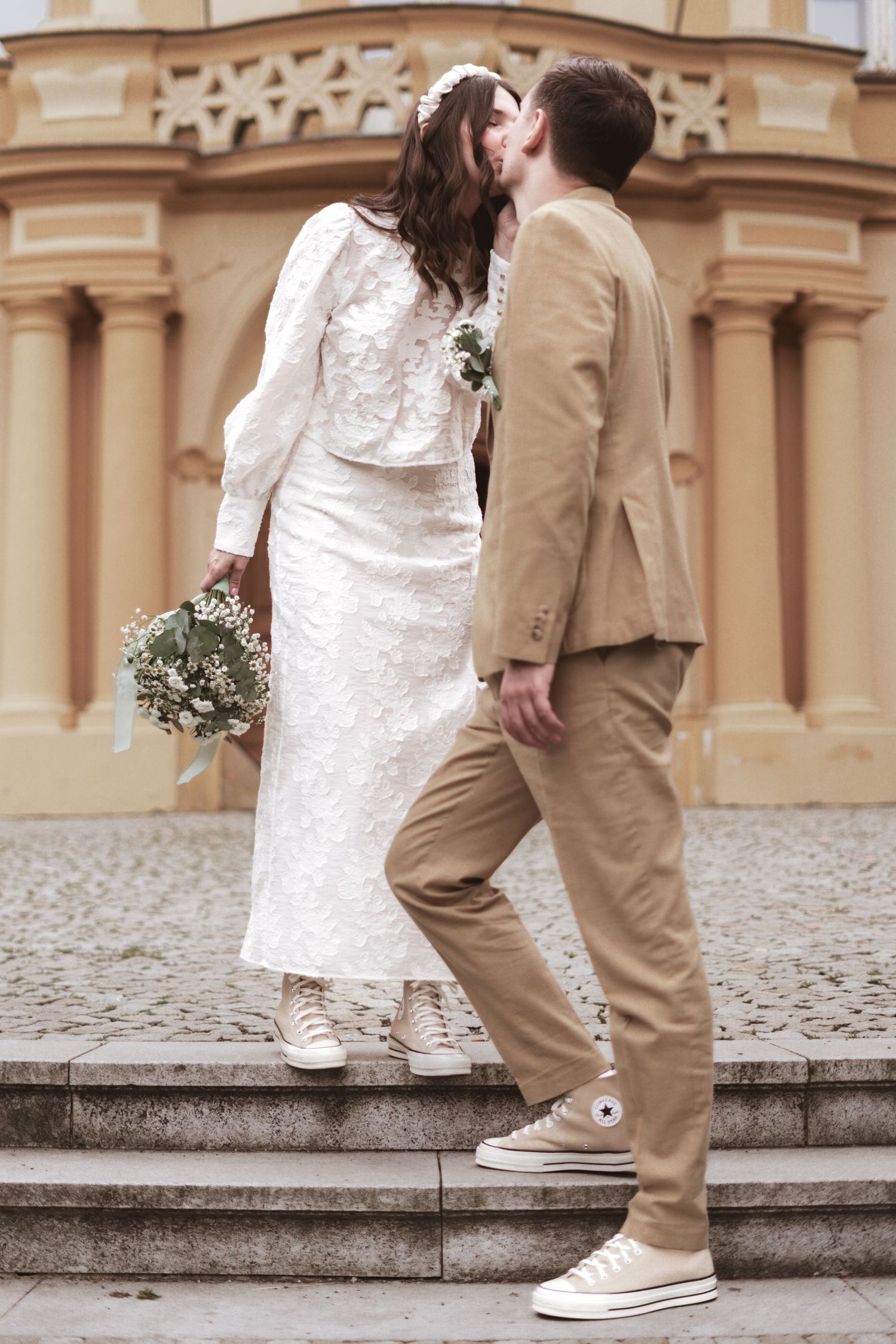 Bride in a white lace outfit and groom in a beige suit kiss on stone steps in front of a historic building, holding a bouquet.