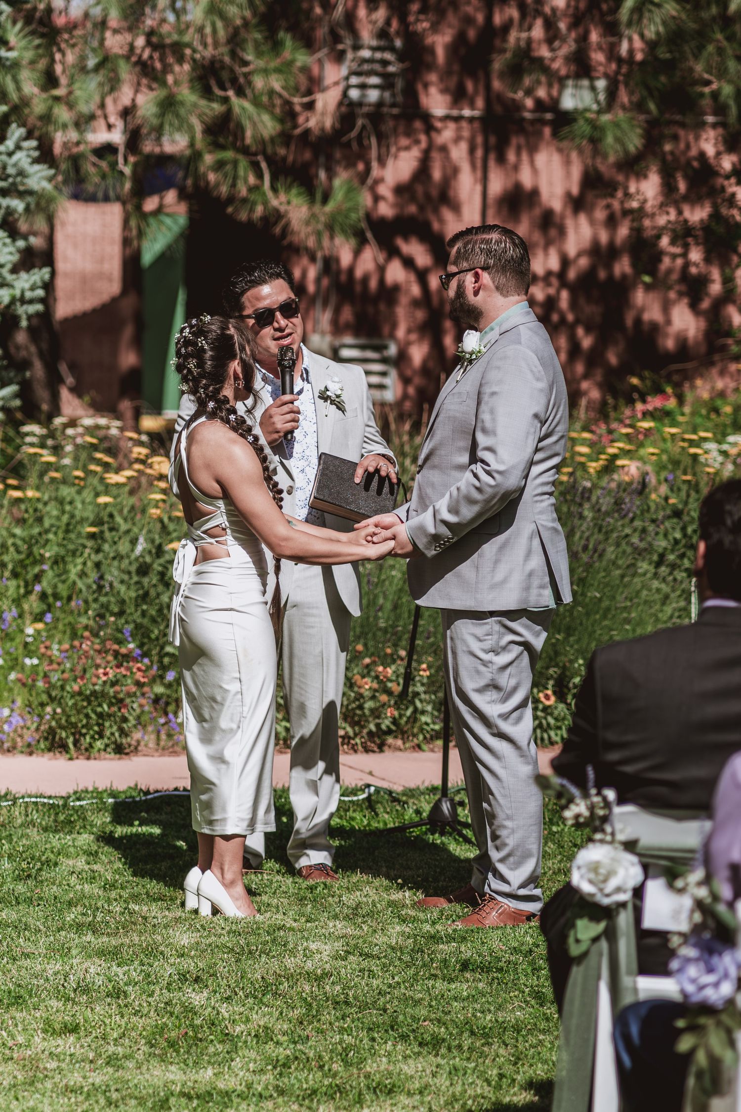 Bride in a white dress and groom in a gray suit hold hands during an outdoor garden wedding ceremony led by an officiant.