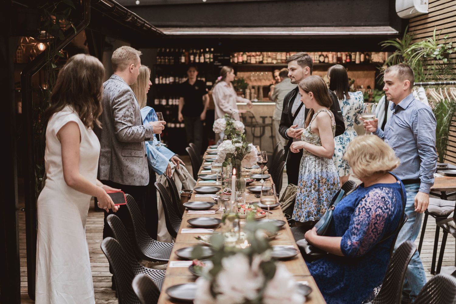 Guests in elegant attire mingle around a long rustic banquet table set with plates and floral centerpieces at a stylish restaurant.