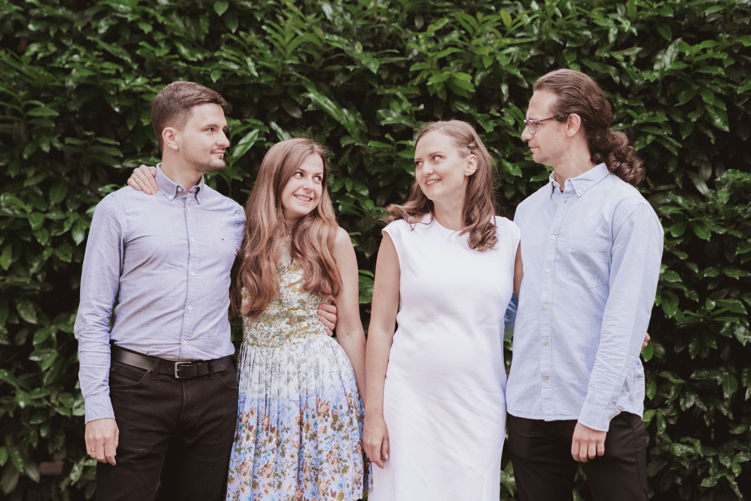 Four young adults, two men and two women, pose arm in arm in front of a lush green hedge, smiling in casual summer outfits.