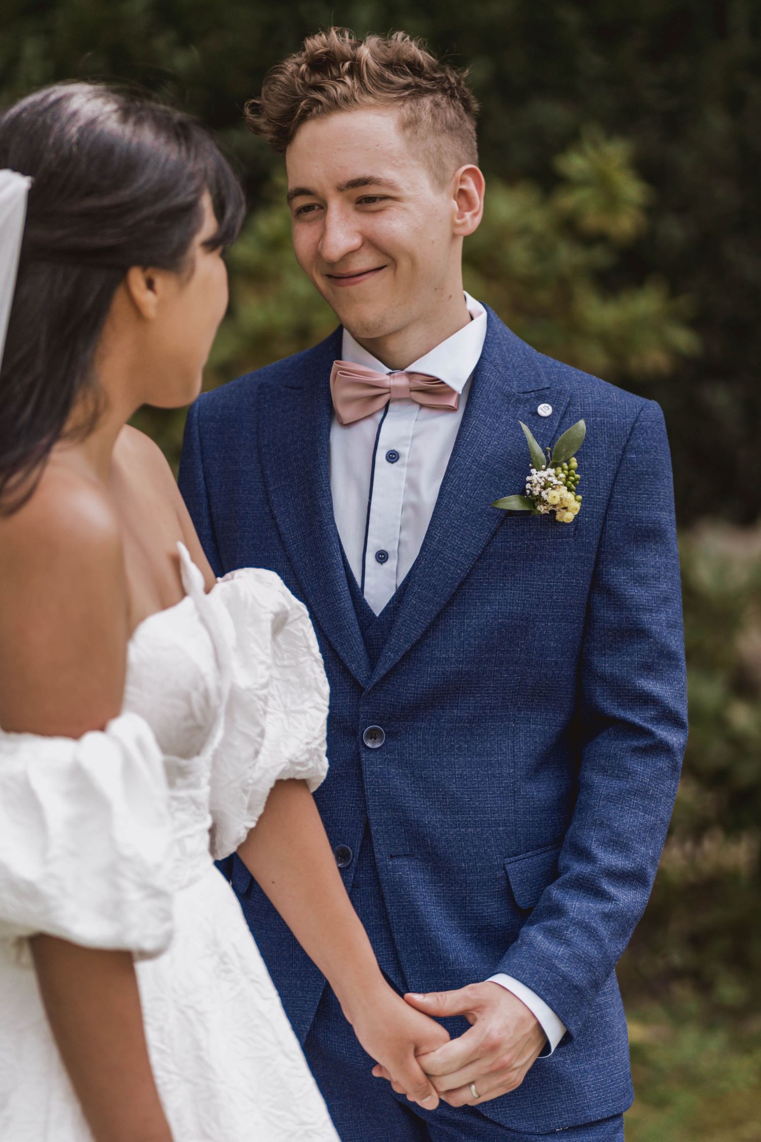 Smiling groom in a blue suit and pink bow tie holds hands with his bride in a white off-the-shoulder gown during an outdoor wedding.