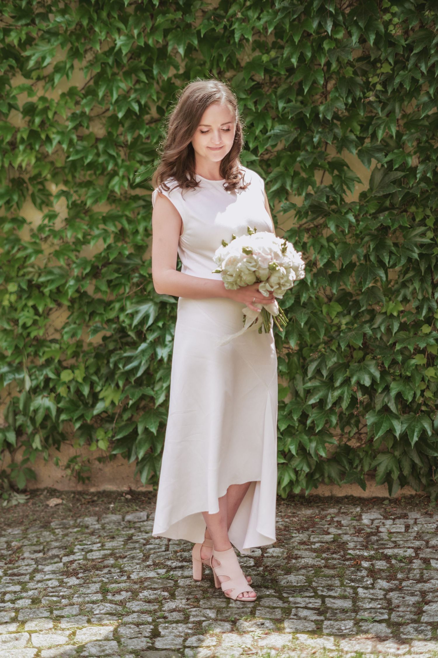 Woman in a sleeveless white midi dress holding a white peony bouquet, standing on cobblestones in front of a green ivy wall.
