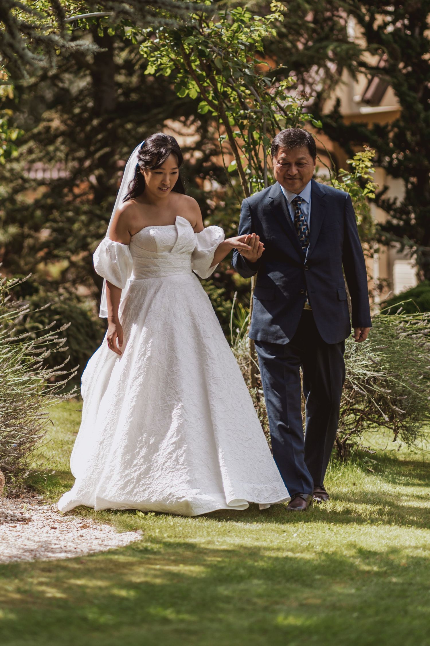 A bride in a strapless white gown with puff sleeves walks arm in arm with a man in a navy suit through a sunlit garden.
