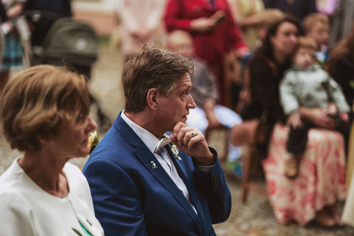 Thoughtful man in a blue suit with a boutonniere listens intently at an outdoor wedding ceremony as guests sit behind him.