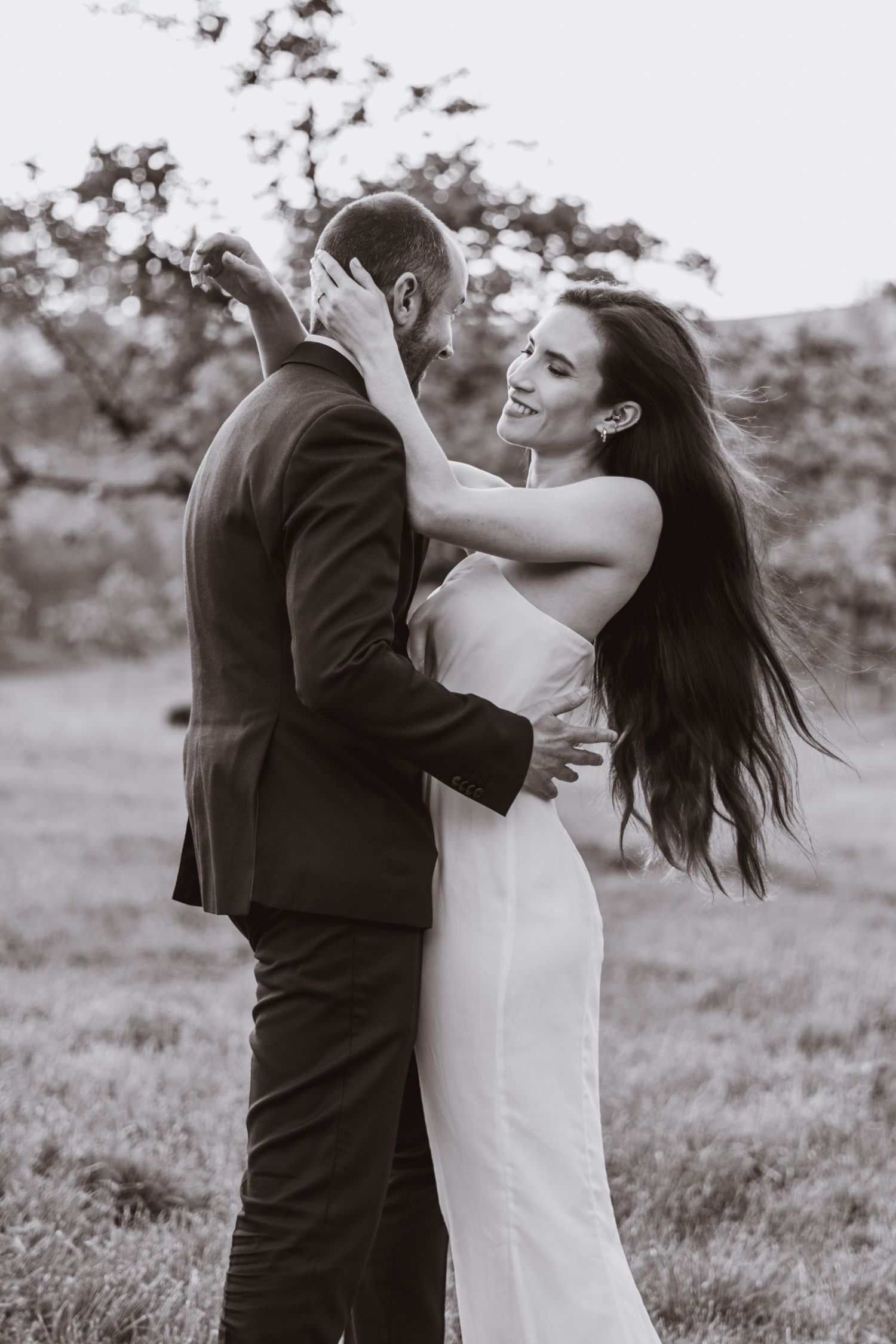 Black and white photo of a couple embracing in a grassy field, the woman in a white dress with long hair blowing in the wind.