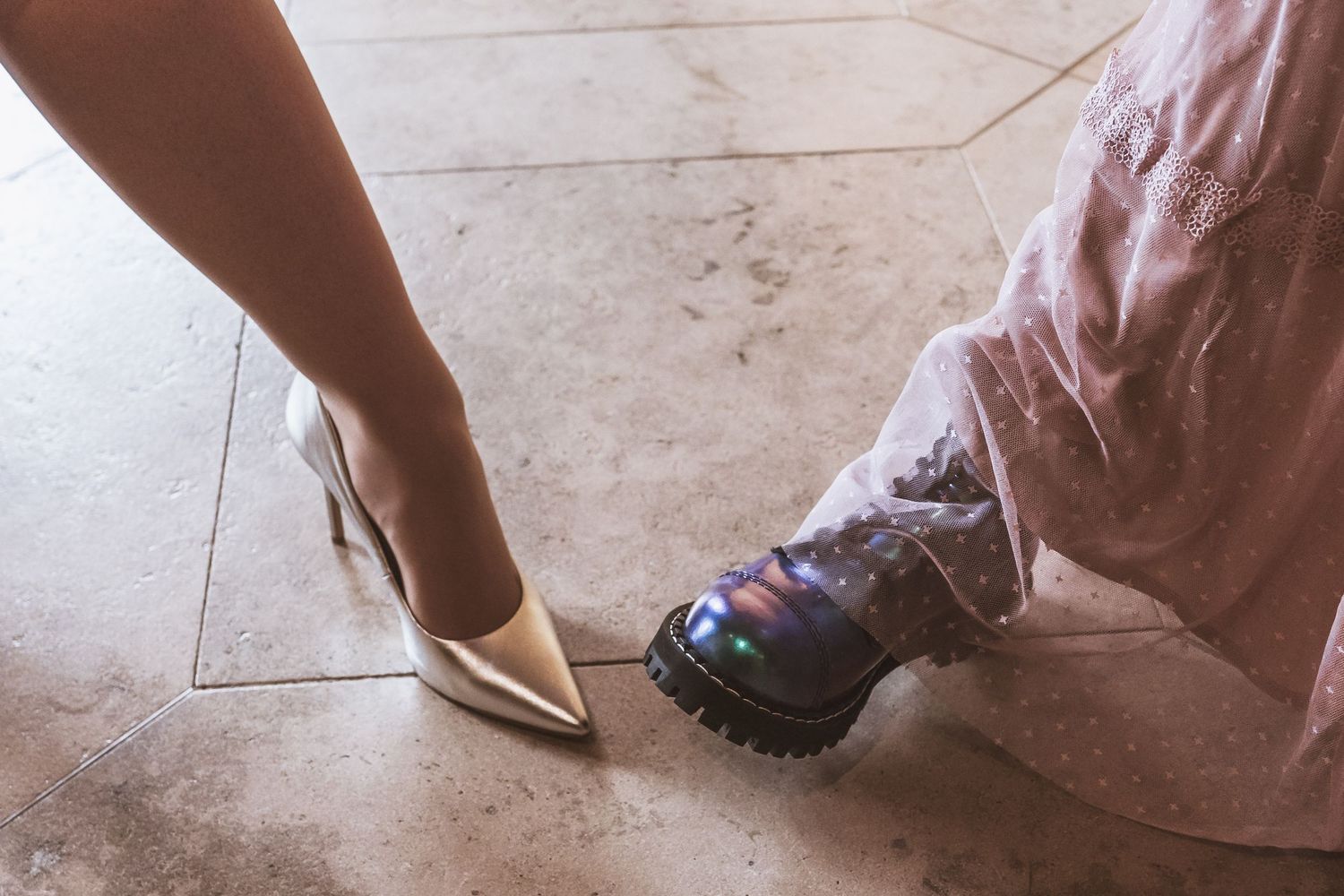 Woman's metallic gold pointed-toe high heel beside a chunky black platform boot with iridescent shine on a light stone tile floor.