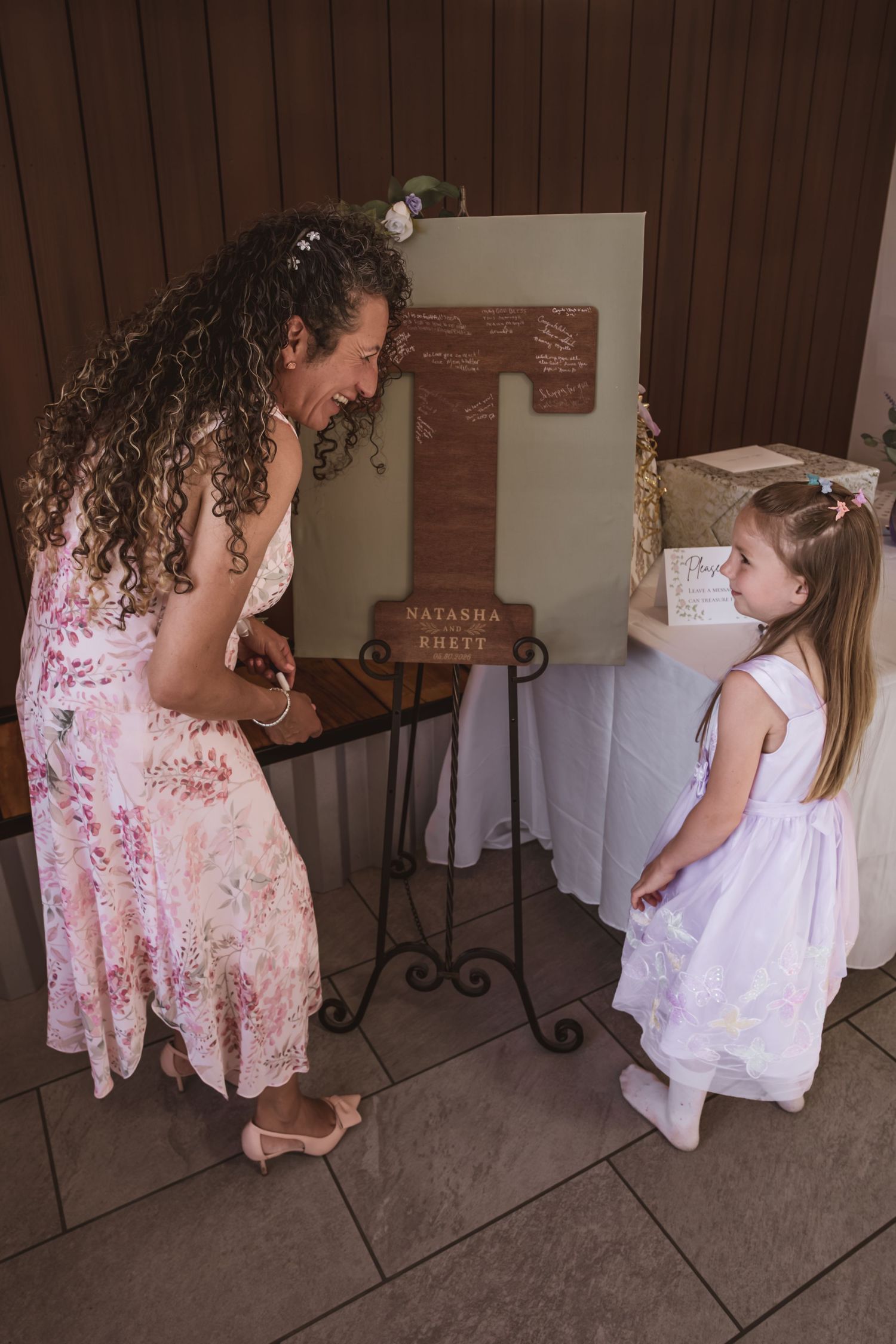 Smiling woman in a floral dress leans toward a young girl in a white dress beside a wooden letter T wedding sign on an easel.