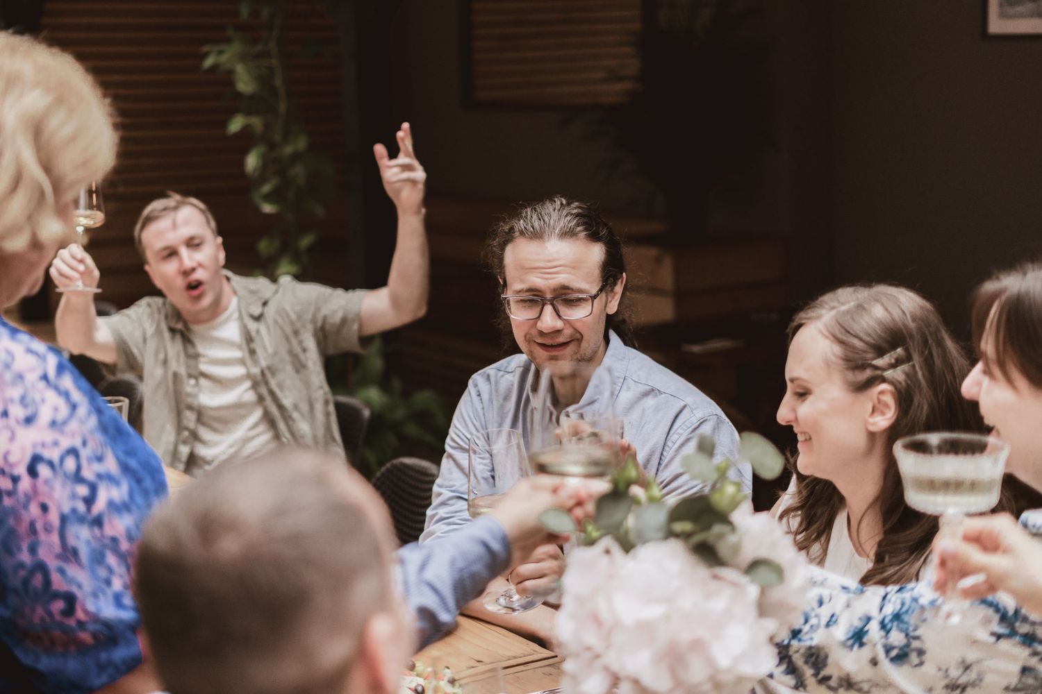 A group of friends laughs and toasts with drinks around a candlelit dinner table, one man raising his hand cheerfully.