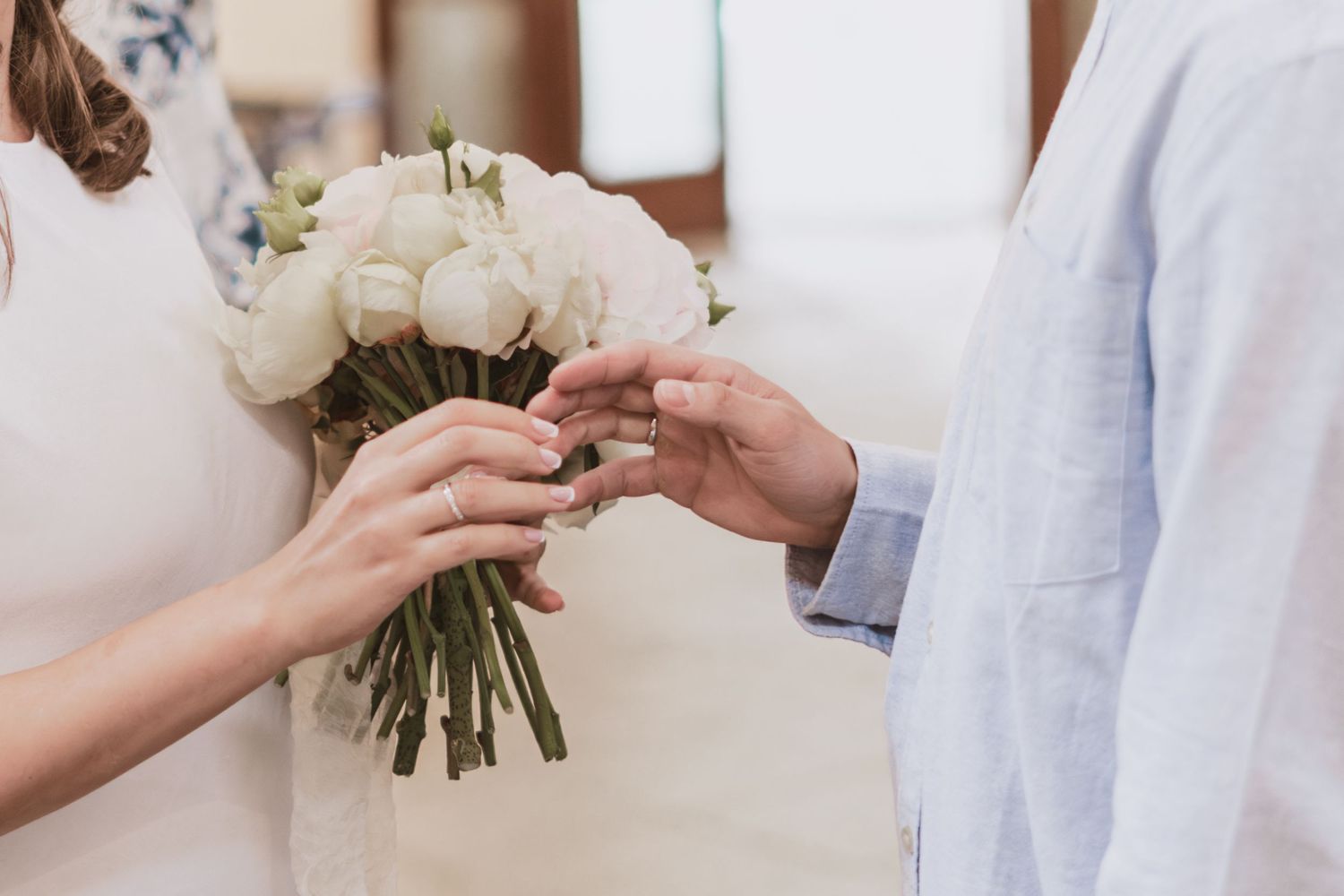 Bride in a white dress holding a bouquet of white roses while a groom in a light blue shirt places a ring on her finger.