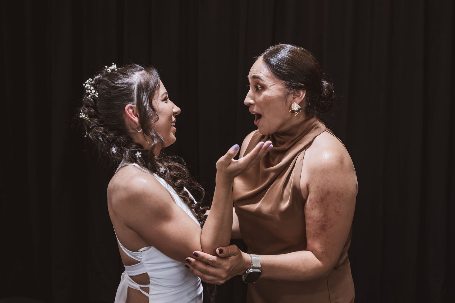 Two women share an emotional, joyful moment, one in a white strappy dress with a floral hairpiece and the other in a satin bronze top.