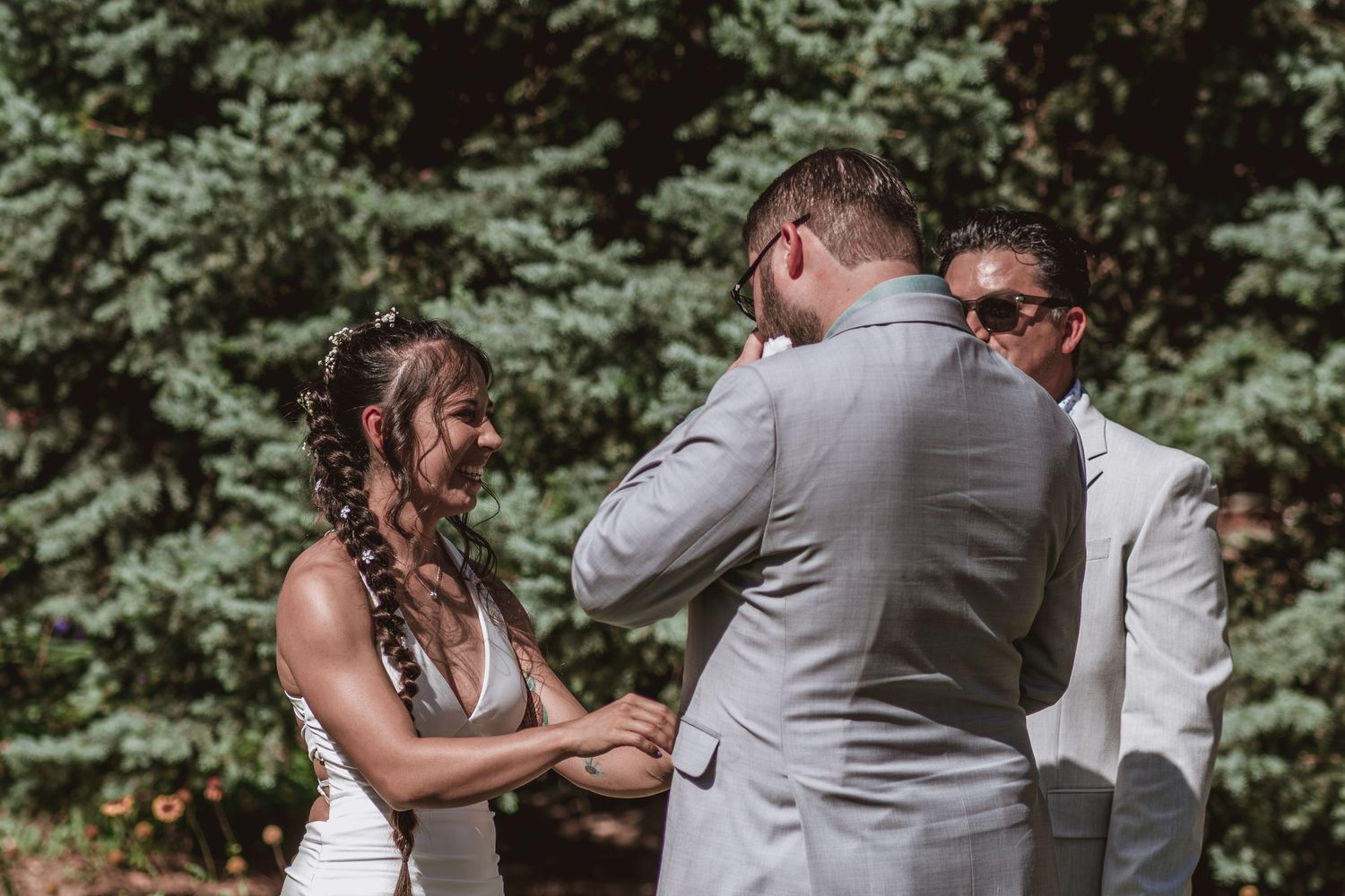 Smiling bride with braided hair holds the groom's hand as he wipes his eyes during an emotional outdoor wedding ceremony.