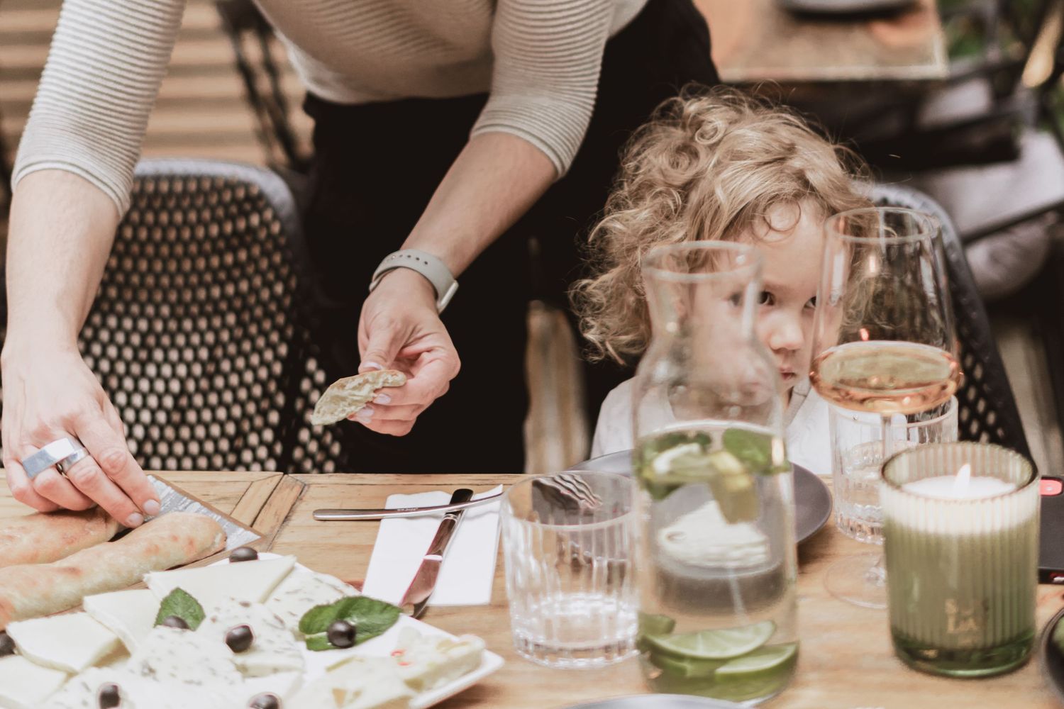 A woman serves cheese and herbs to a curly-haired child at a restaurant table with lemon water, a rosé glass and a lit candle.