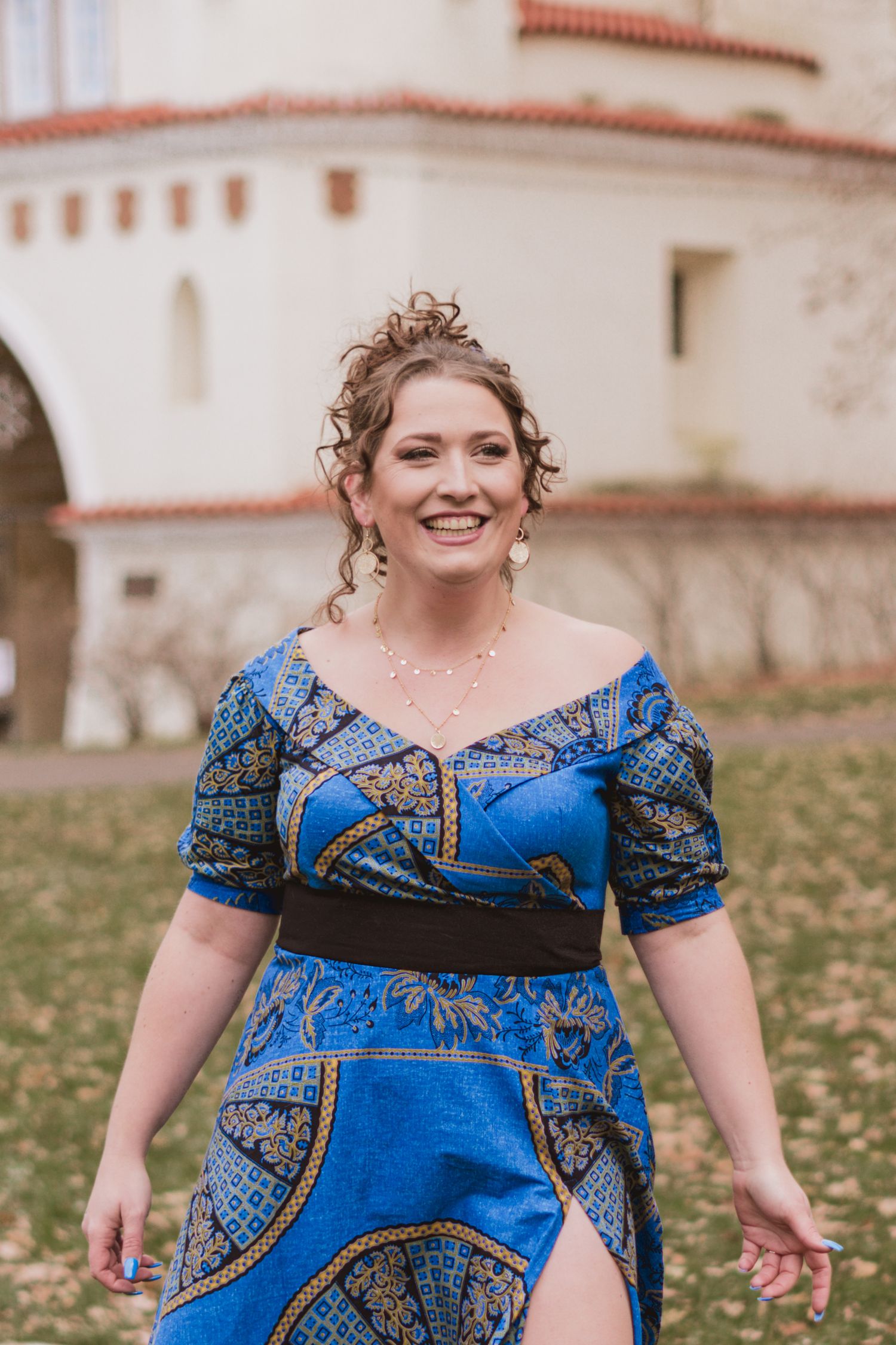 Smiling woman with an updo walks across a leaf-strewn lawn in a blue patterned African print dress with a black belt and side slit.