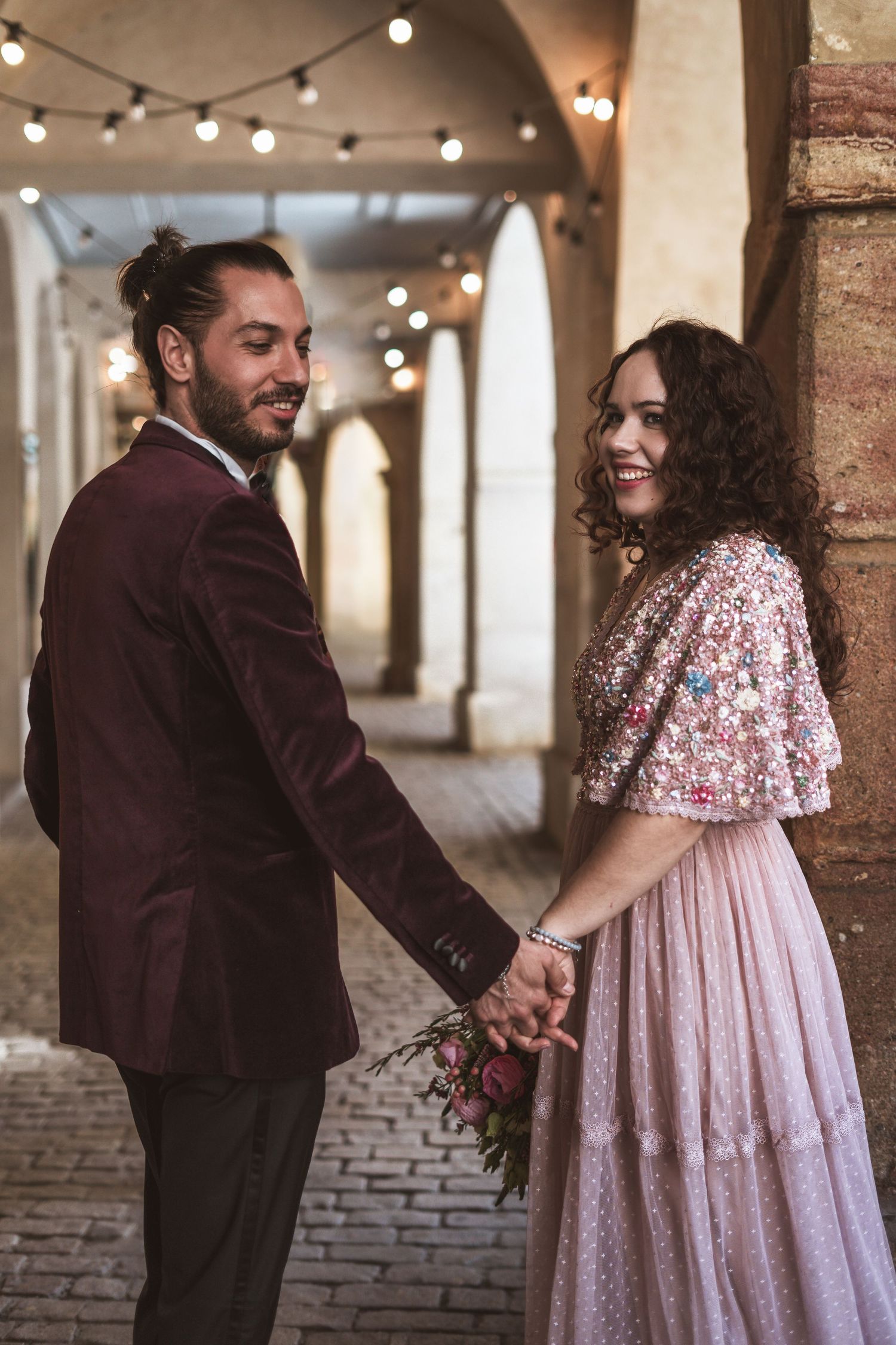 Smiling couple holding hands under a stone arcade with string lights, the man in a burgundy velvet blazer and the woman in a pink floral dre