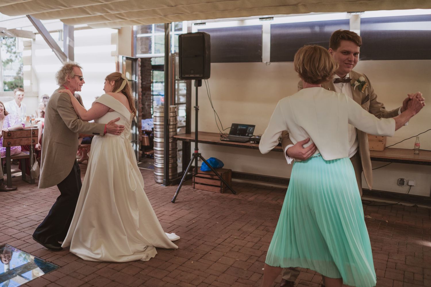 Bride in a white gown dances with an older man while a couple in mint green skirt and tan jacket dances nearby at a wedding reception.