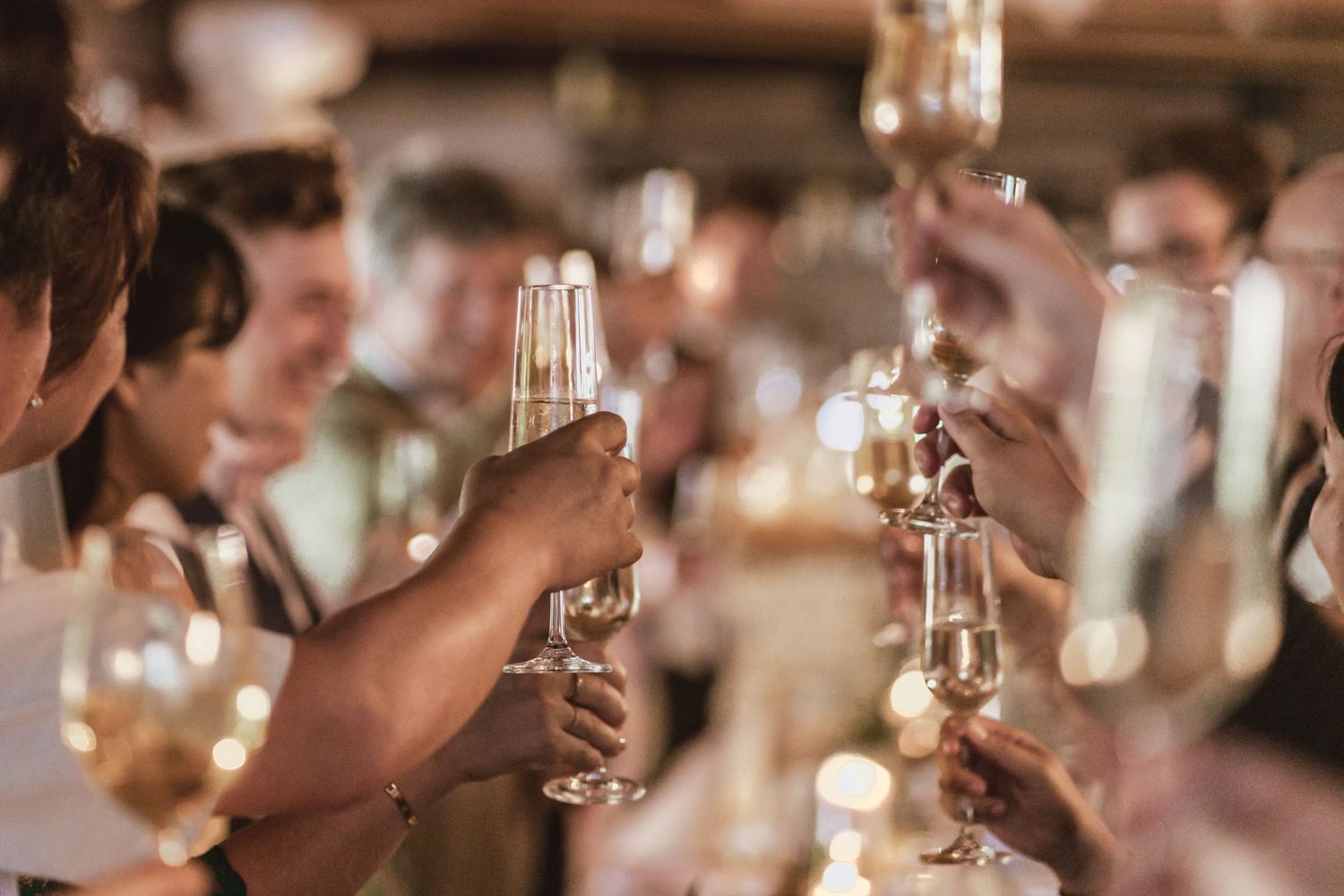 Group of friends raising glasses of sparkling wine in a lively toast at a festive celebration with warm, glowing lights.