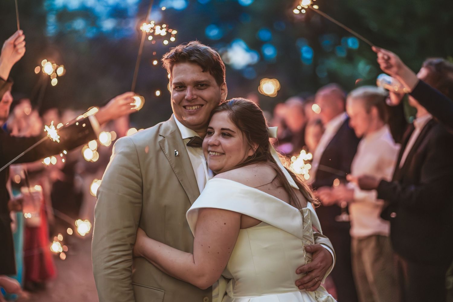 Smiling bride and groom embrace at a evening wedding reception while guests hold glowing sparklers in the background.