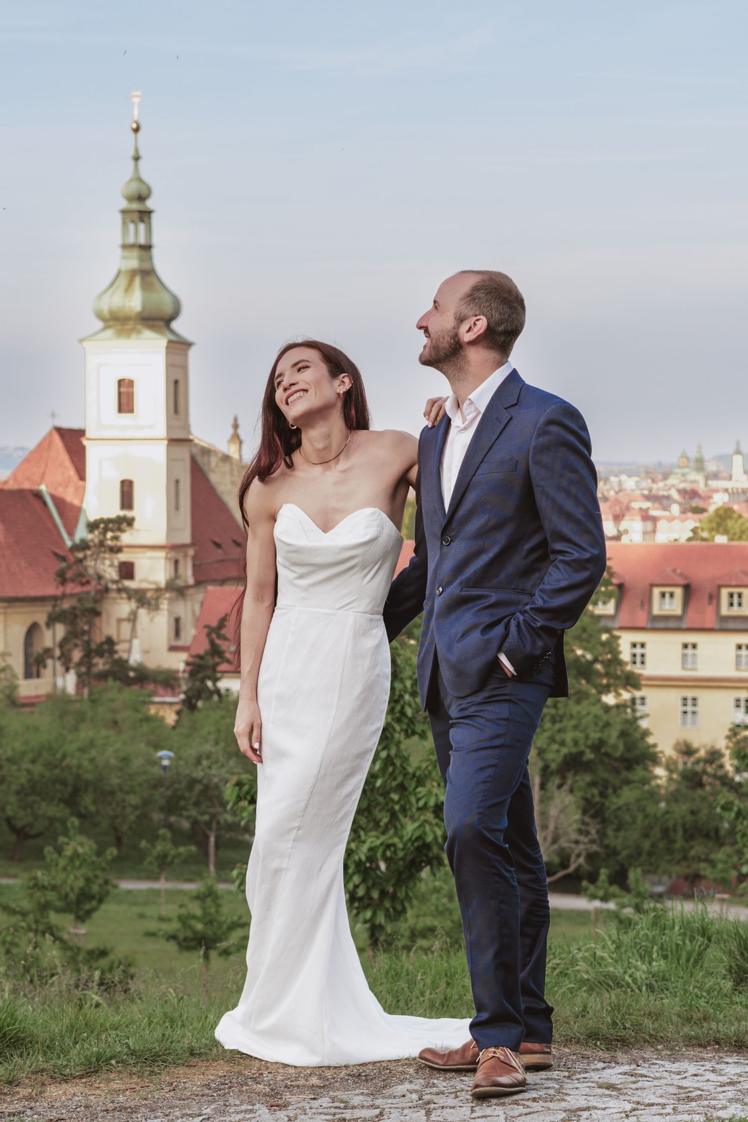 Bride in a white strapless gown and groom in a navy suit smile and look up together on a hill overlooking a historic European town.
