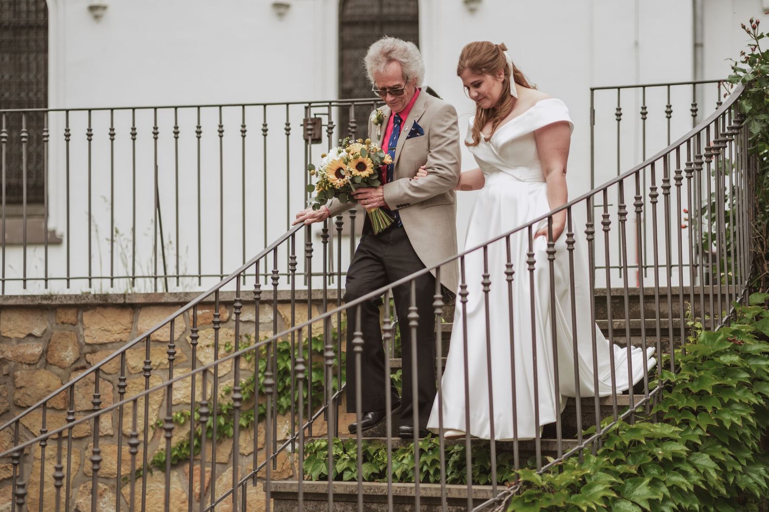 A father in a gray suit escorts a bride in a white gown down an ornate wrought iron staircase, holding a bouquet of yellow flowers.