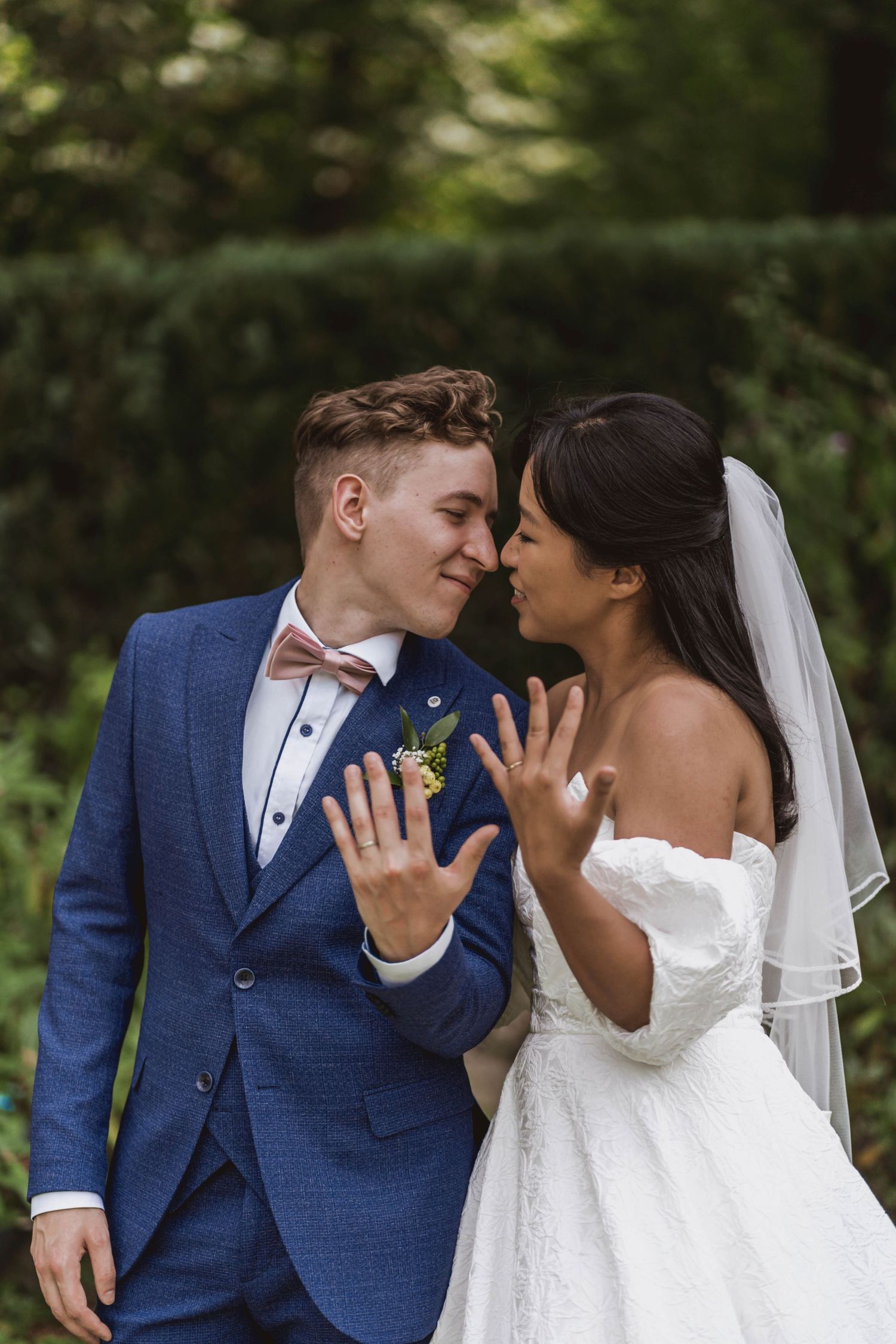Smiling bride and groom touch noses outdoors while showing their wedding rings, surrounded by green hedges.