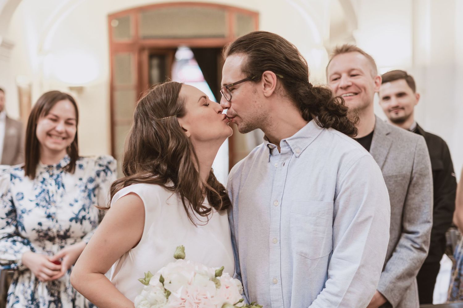 A bride in a white dress and a long-haired groom with glasses share a kiss while smiling wedding guests look on.
