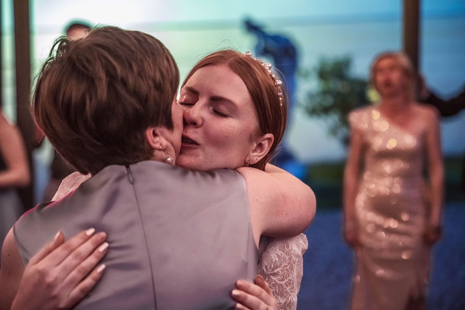 Two women share a warm, emotional hug at a wedding reception, one wearing a bridal hair accessory, with a guest in a sequined dress behind.