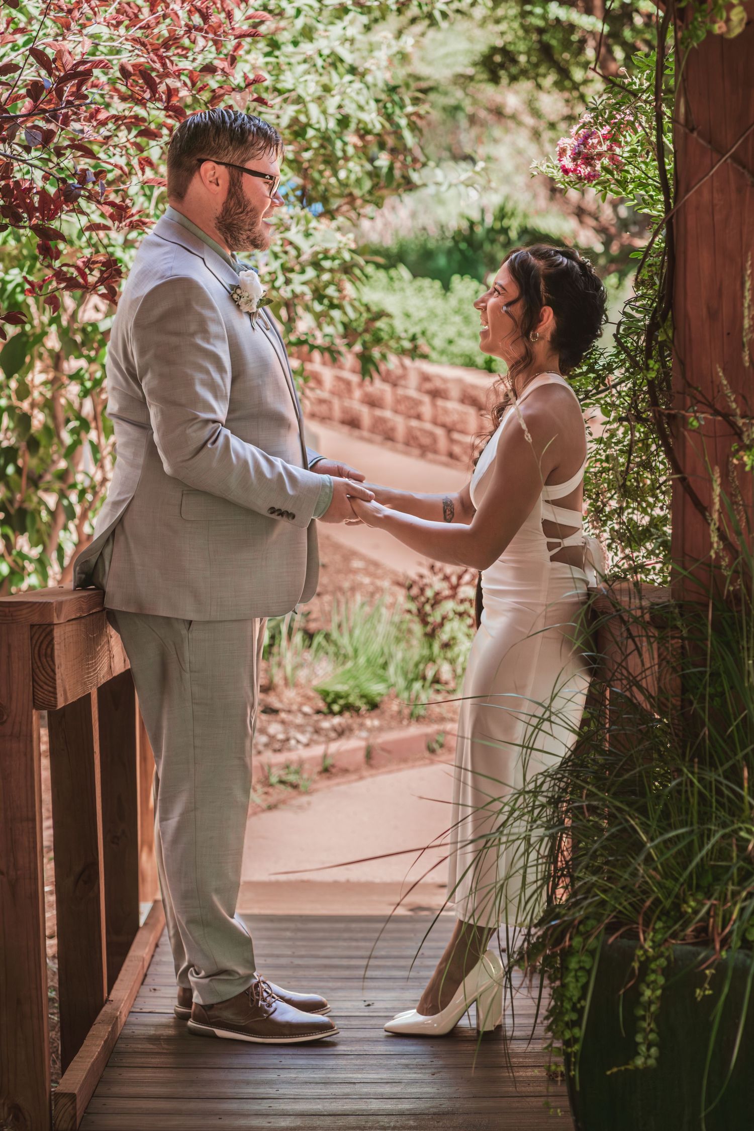 Bride and groom hold hands facing each other on a wooden garden bridge, surrounded by lush greenery at an outdoor wedding ceremony.