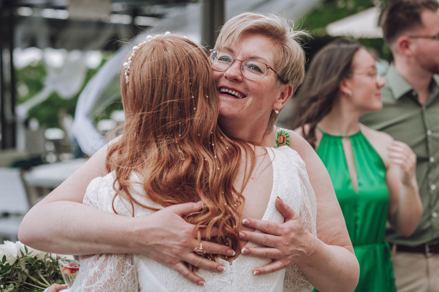 A smiling older woman with glasses embraces a red-haired bride in a white lace dress during a joyful outdoor wedding celebration.