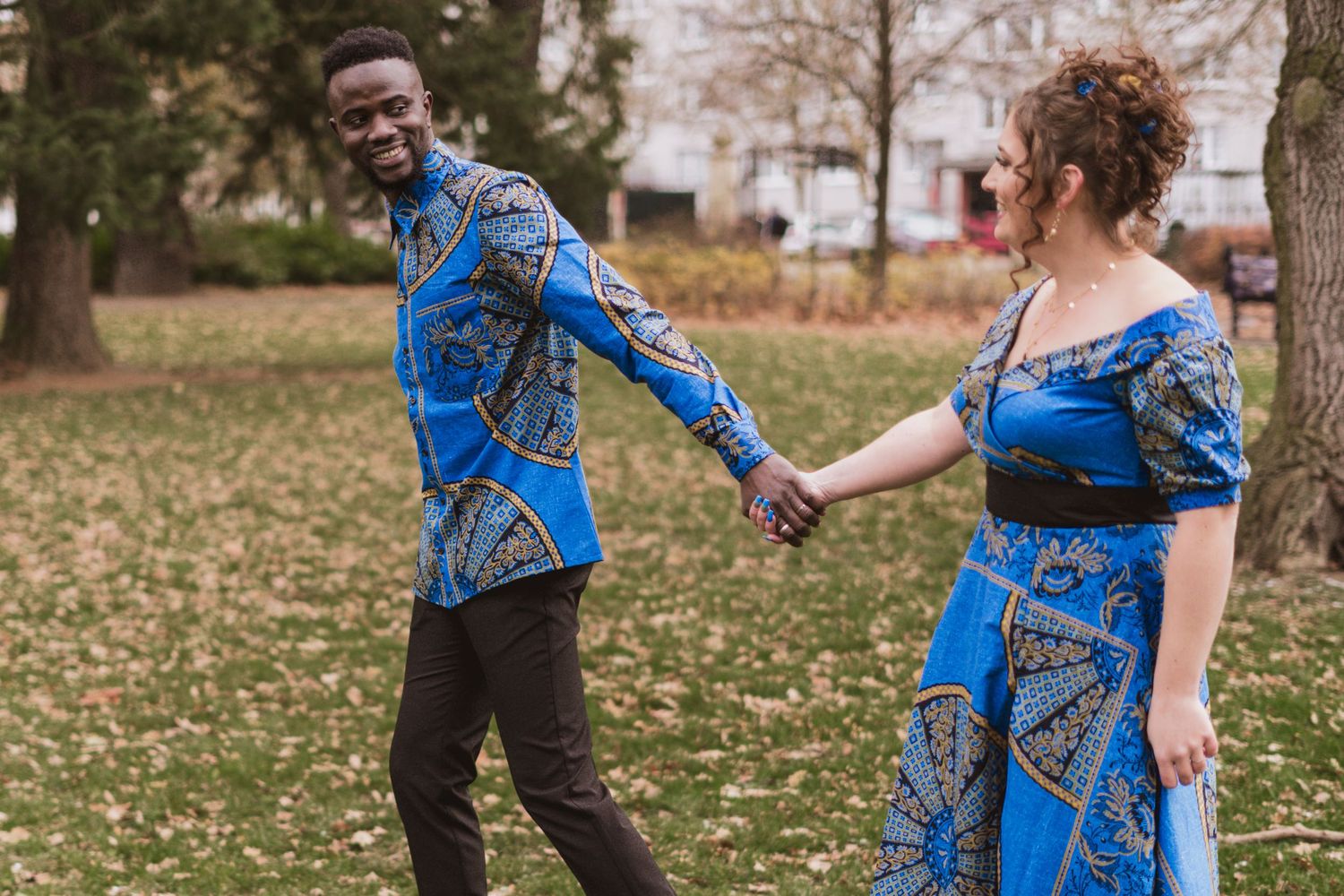 Smiling couple in matching blue African print outfits holding hands while walking across a sunny park lawn with autumn trees.