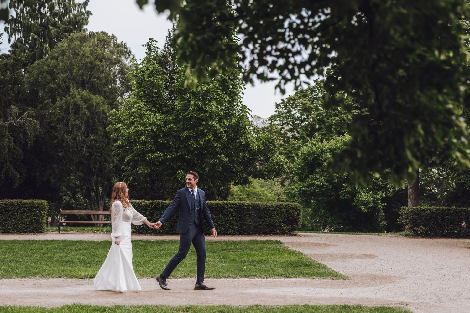 A bride in a white gown and a groom in a navy suit walk hand in hand along a path in a leafy green park.