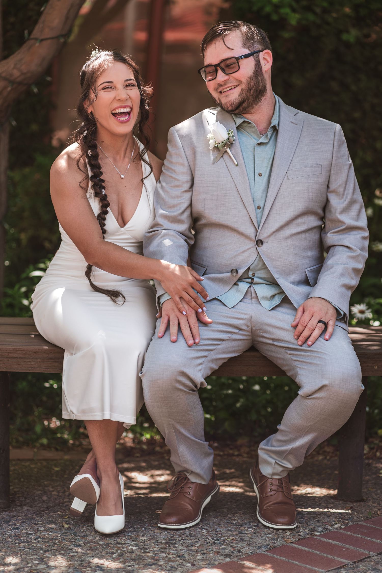 A laughing bride in a white dress and a bearded groom in a gray suit and glasses sit close together on a wooden bench outdoors.