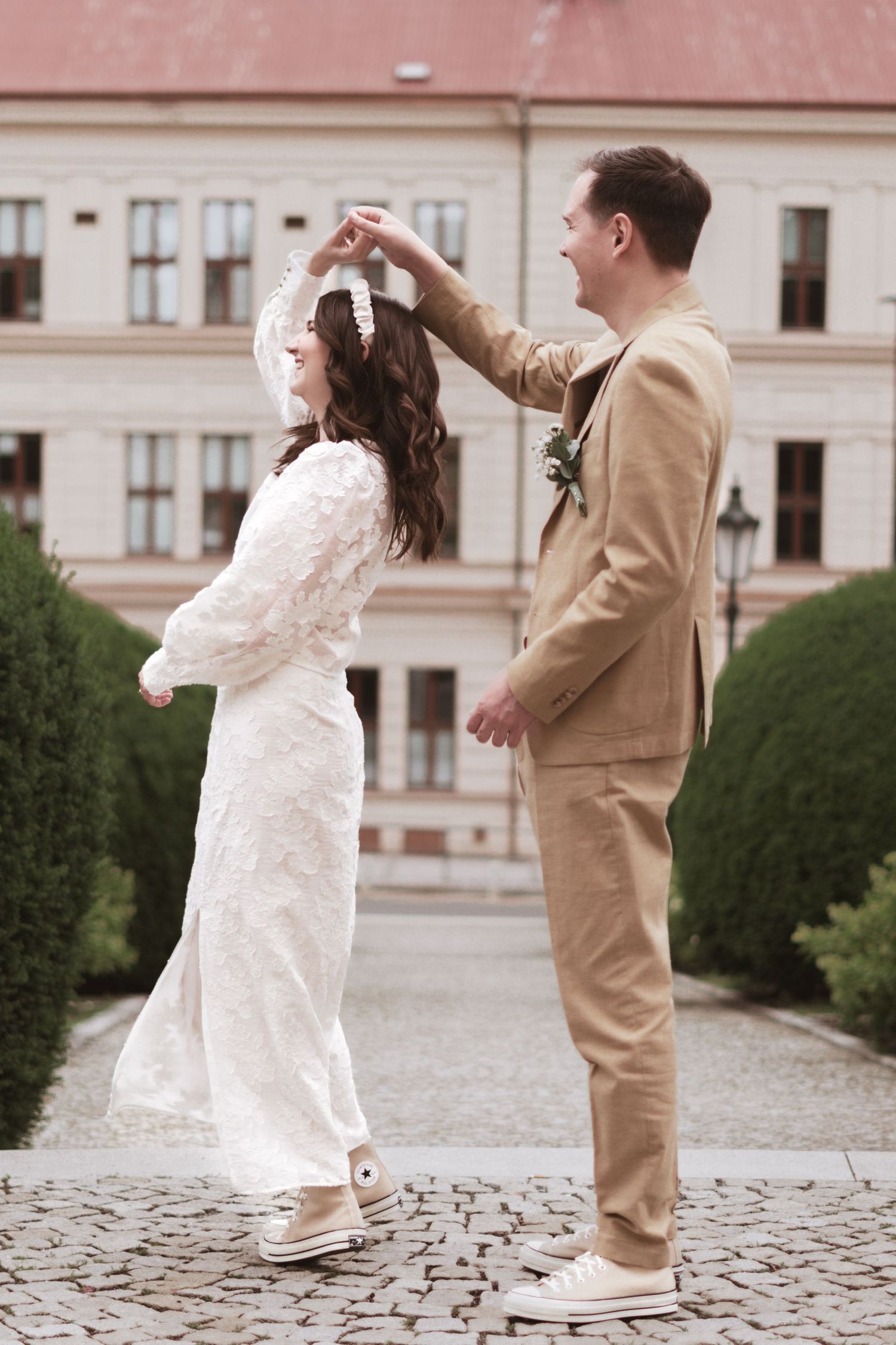 A bride in a lace dress twirls under her groom's raised hand as they dance on a cobblestone path outside a historic building.