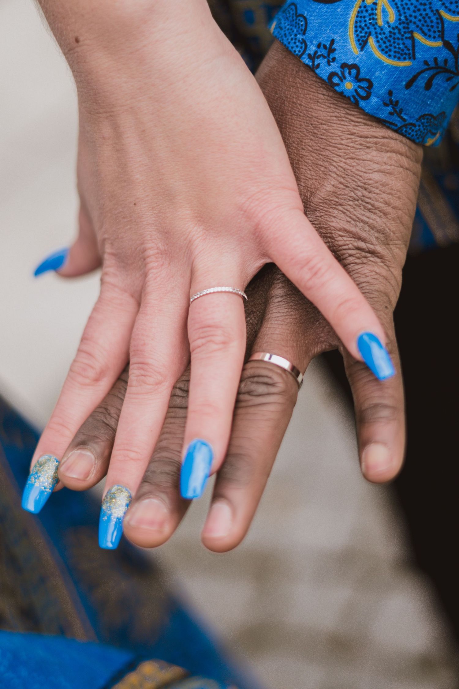 Close-up of two hands resting together showing silver wedding rings and blue manicured nails, beside a blue patterned sleeve.