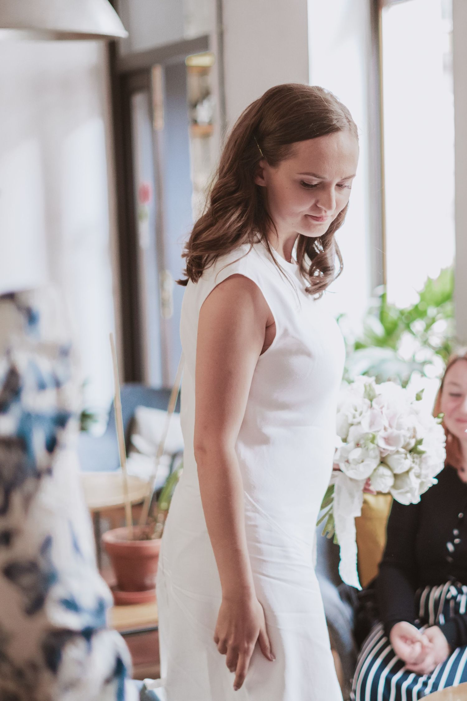 Smiling woman in a sleeveless white dress looks down in a bright room while another woman holds a bouquet of white flowers nearby.