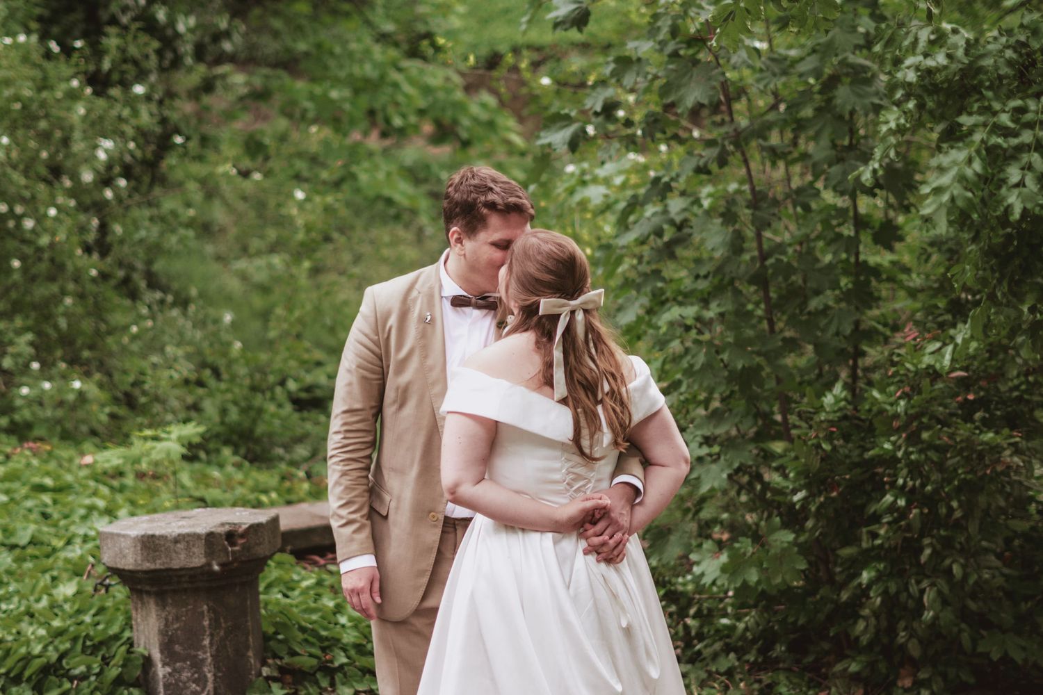 Bride in a white off-the-shoulder gown embraces a groom in a tan suit and bow tie in a lush green garden beside a stone pedestal.