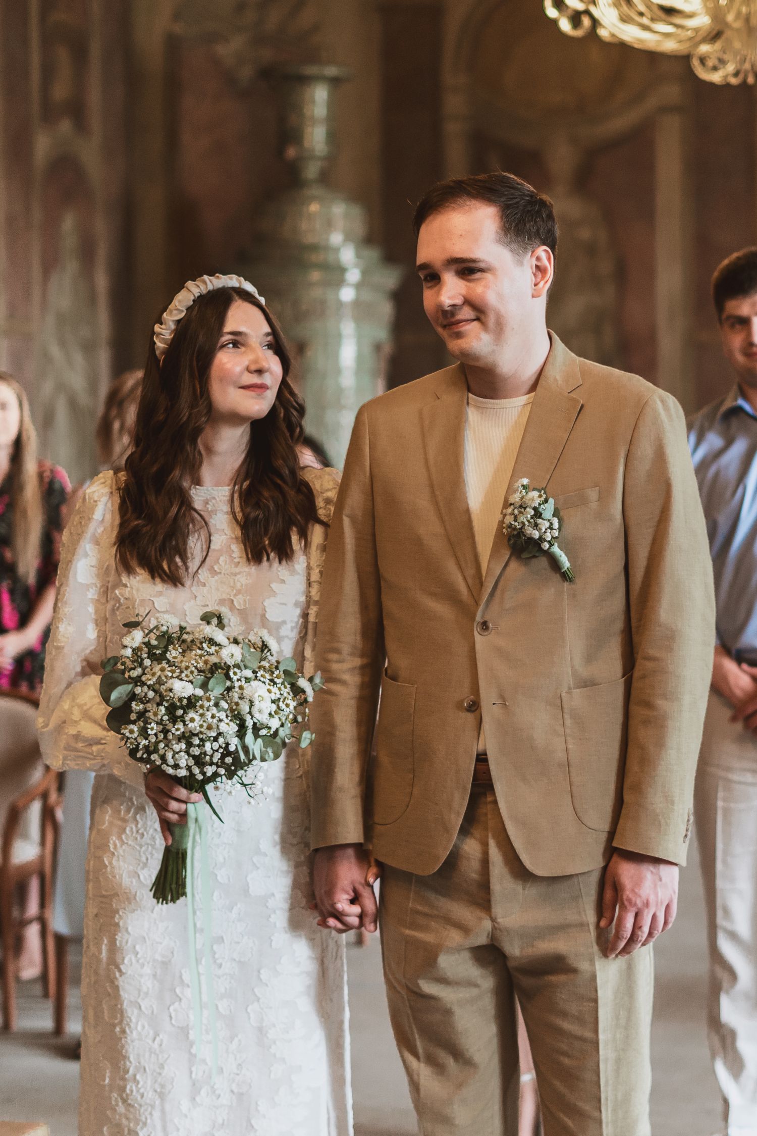Smiling bride in a lace dress and floral headband holds hands with a groom in a tan suit during a wedding ceremony.