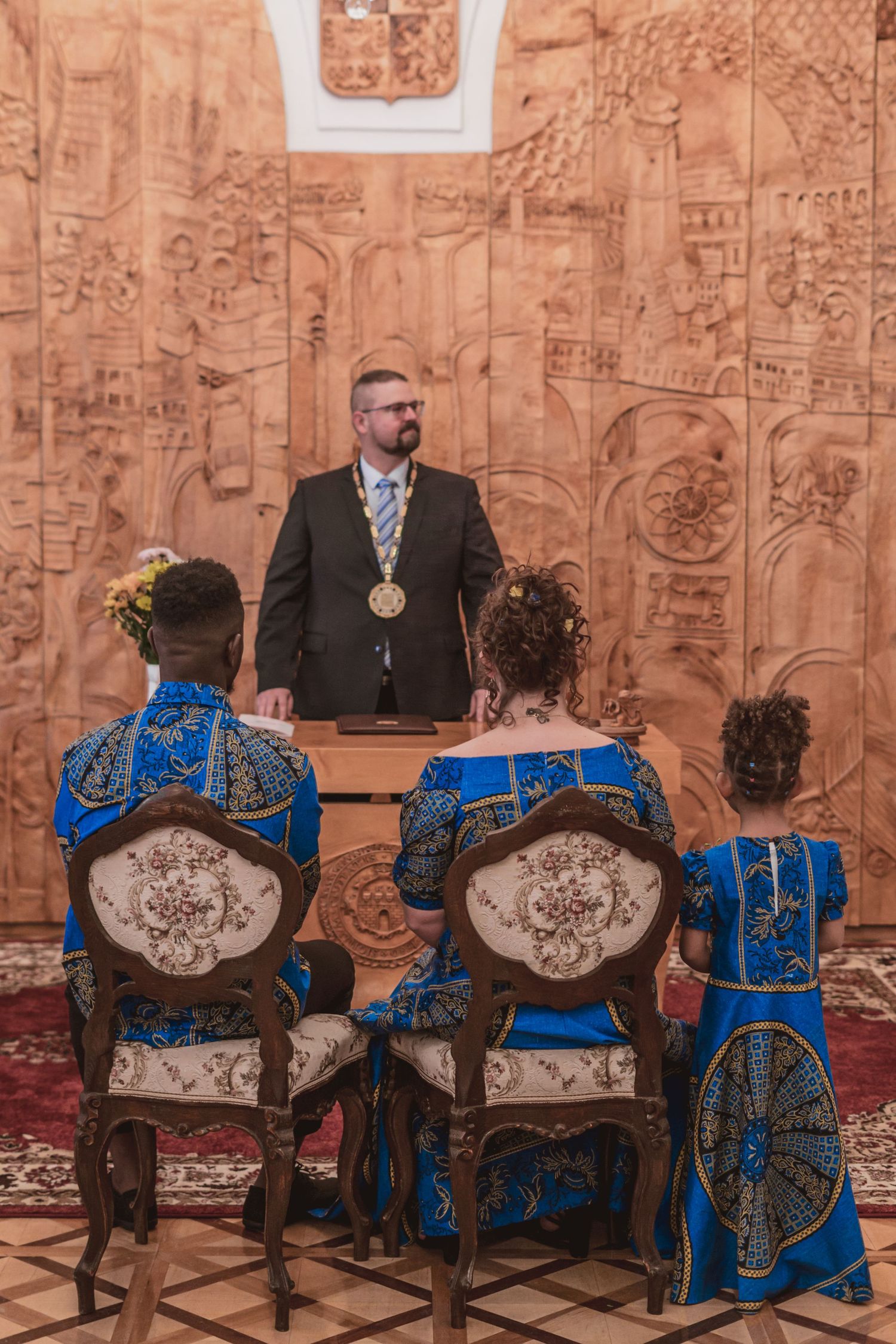 Couple in matching blue African print outfits and a young girl sit before an official wearing a medal chain during a civil wedding ceremony.