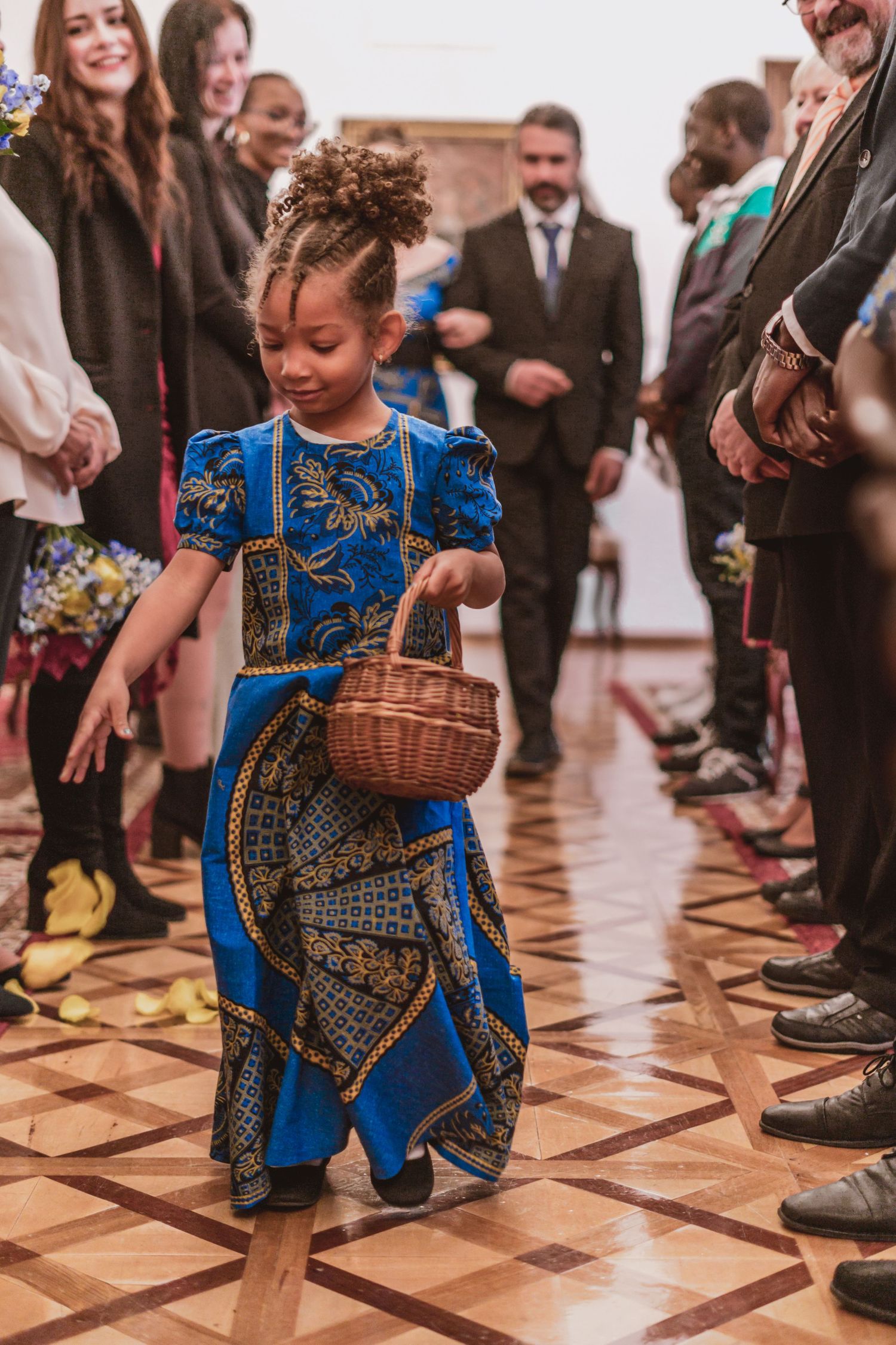 Young flower girl in a blue African print dress carries a wicker basket down an aisle lined with smiling wedding guests.