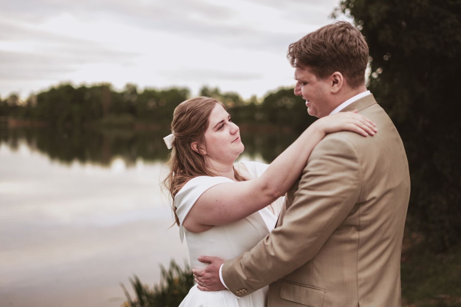 A bride in a white dress and a groom in a tan suit embrace in a tender moment beside a calm lake at sunset.