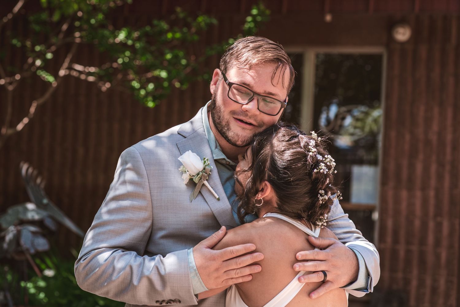 Smiling groom in a gray suit and floral boutonniere embraces a bride in an updo with flower hairpins during an emotional wedding moment.