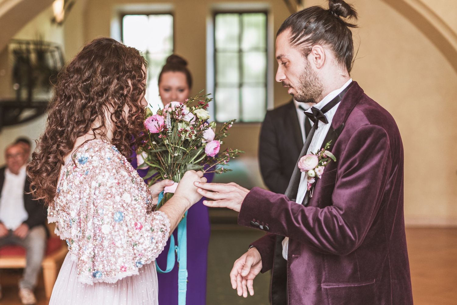Groom in a burgundy velvet suit and bow tie reaches toward a bride holding a pink bouquet during a rustic indoor wedding ceremony.