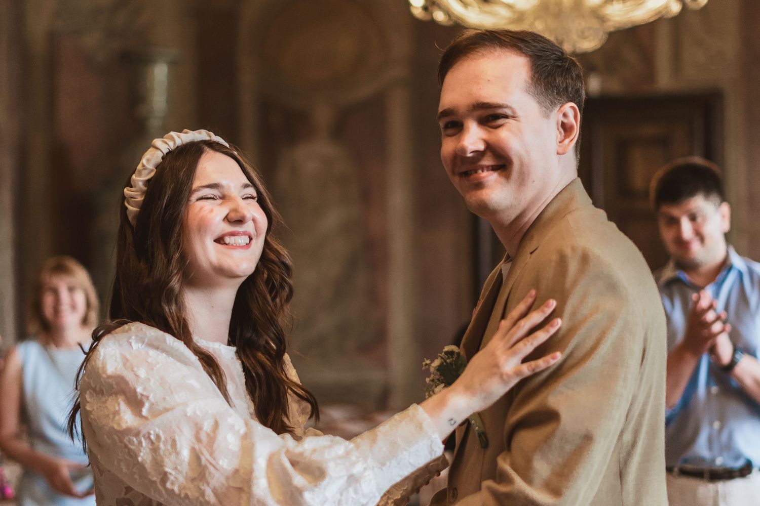 Smiling bride in a white lace dress and floral headband laughs with her groom in a tan suit during a wedding ceremony as guests look on.