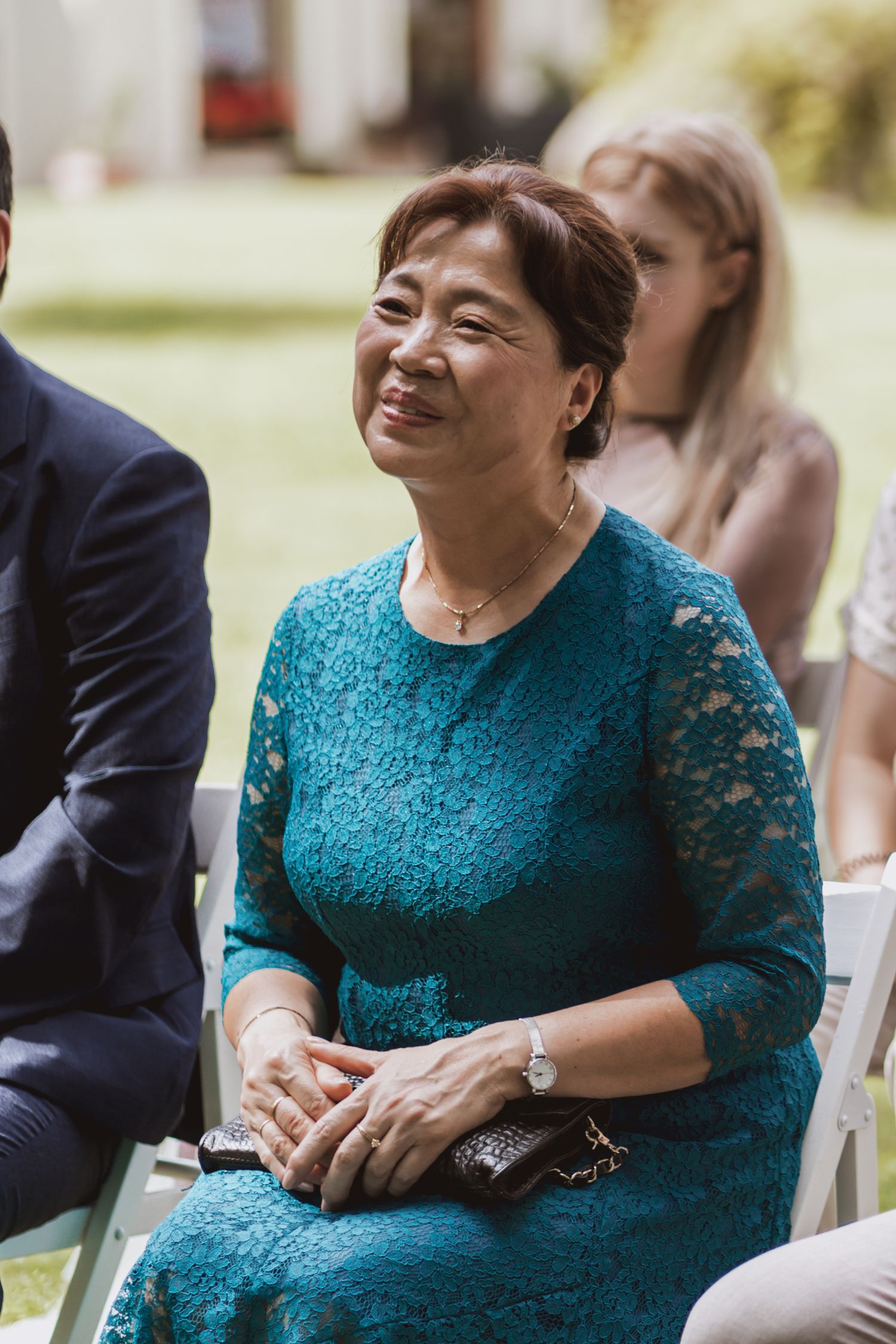 Smiling woman in a teal lace dress sits with hands folded on her lap among guests at an outdoor wedding ceremony.